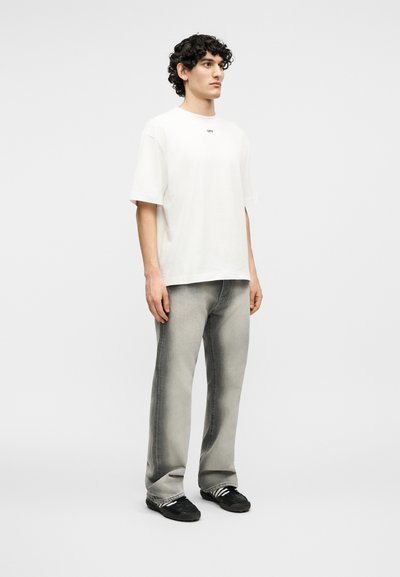 Νέος άνδρας με σγουρά μαλλιά στέκεται, φορώντας ένα λευκό υπερμεγέθες t-shirt, ξεθωριασμένο γκρι τζιν και μαύρα αθλητικά παπούτσια με άσπρες ρίγες.