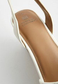 Sandale en cuir blanc avec une semelle intérieure rembourrée couleur tan. Présente des bords cousus et un logo embossé sur la semelle intérieure. Conception à bout ouvert et fines lanières.