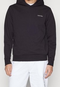 Man in een zwarte Calvin Klein hoodie en witte broek met het Calvin Klein logo op de linkerdij.
