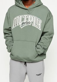 Sudadera con capucha verde que presenta un gran gráfico de texto blanco que dice "MULTIPLY" con un acento en cursiva. Combinada con pantalones de chándal grises.