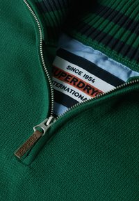 Grüner Strickpullover mit Reißverschluss, ausgestattet mit einem gerippten Kragen in Dunkelgrün und Marineblau. Das Etikett trägt die Aufschrift "Superdry seit 1954."