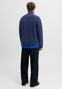 Suéter de punto azul marino con cuello alto, dobladillo acanalado, usado sobre una blusa azul, combinado con pantalones negros a rayas y zapatos negros. Vista trasera.
