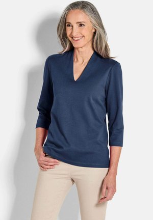 Navyblaues V-Ausschnitt-Oberteil mit Dreiviertelärmeln, gefertigt aus einem glatten, dehnbaren Stoff. Helle beige Hosen ergänzen das Outfit.