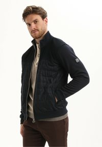 Marineblauwe zip-up jas met geribbelde schouders en een gewatteerd lijf. Heeft twee voorzakken en een geribbelde zoom. Daaronder zit een beige henley shirt.