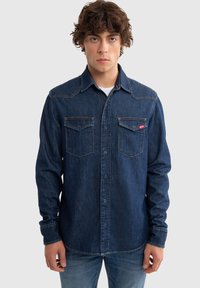 Camisa de mezclilla azul oscuro con botones a presión, dos bolsillos en el pecho y costuras en contraste. Presenta un cuello puntiagudo y mangas largas.