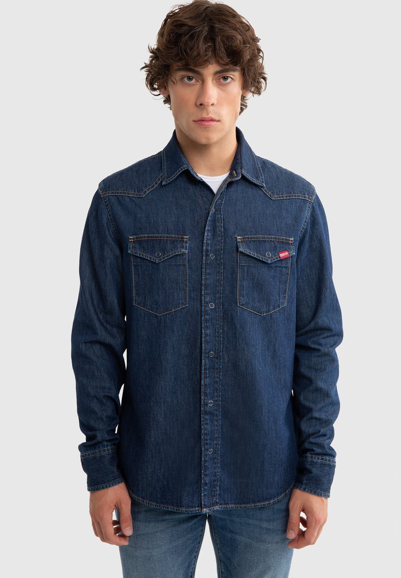 Camisa de mezclilla azul oscuro con botones a presión, dos bolsillos en el pecho y costuras en contraste. Presenta un cuello puntiagudo y mangas largas.