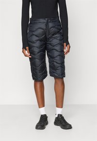 Calções pretos acolchoados com um design acolchoado, apresentando uma cintura alta e fecho frontal. Usados com sapatos pretos e meias brancas.