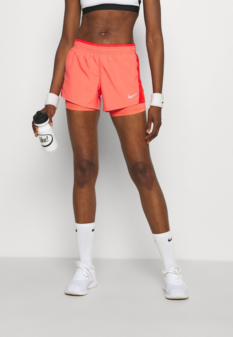 Pantalones cortos deportivos Nike en color coral brillante, material ligero, cintura elástica, combinados con zapatillas y calcetas blancas, sosteniendo una botella de agua blanca.