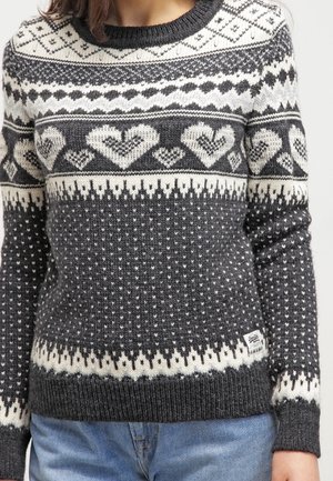 Femme portant un pull tricoté gris foncé et blanc avec des motifs de cœur et géométriques, associé à un jean bleu, montrée du cou aux hanches.
