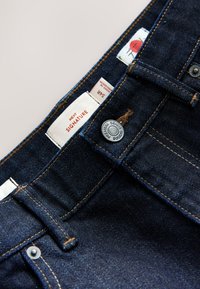 Donkerblauwe spijkerbroeken in close-up met een metalen knoop waarop "Premium Denim Goods" staat en stoffen labels met maat- en merkdetails.