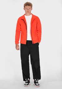 Heldere oranje zip-up jack met een hoge kraag, gecombineerd met losse zwarte broek en zwart-witte sneakers. Glad textuur overal.