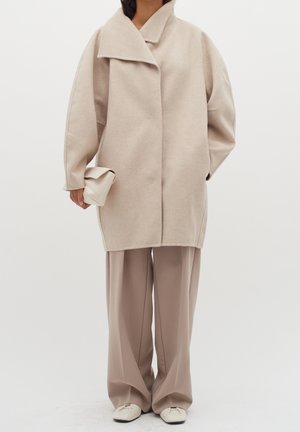 Person iført en oversized beige frakke, vidde taupe bukser, hvide sko med firkantet snude og holder en foldet beige clutchtaske.