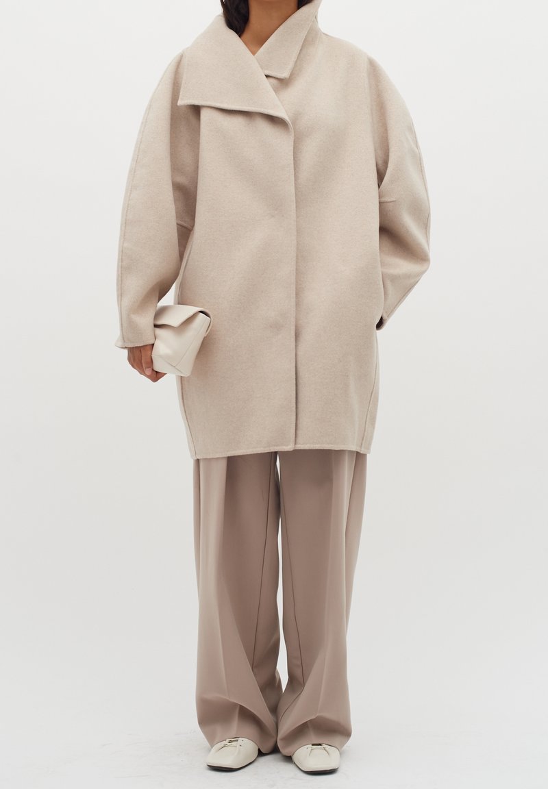 Personne portant un manteau beige oversize, un pantalon large taupe, des chaussures blanches à bout carré, tenant une pochette beige pliée.