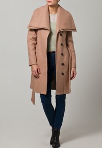 Beige ullrock med en vid krage, dubbelknäppt design och sex svarta knappar. Bärs över en ljusgrön tröja och blå jeans.