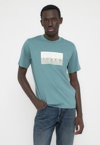 Μπλουζάκι από βαμβάκι σε χρώμα τιρκουάζ με στρογγυλή λαιμόκοψη, με λευκή ορθογώνια σχεδίαση λογότυπου που γράφει "JACK & JONES" με γκρίζα γράμματα, συνδυασμένο με μπλε τζιν παντελόνι.