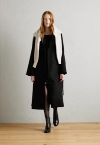 Lange zwarte jas met een witte faux fur kraag en zijsplitten, gecombineerd met een zwarte jurk en enkellaarsjes, tegen een neutrale achtergrond.
