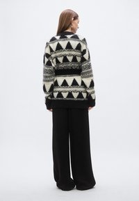 Πλεκτή ζακέτα σε μαύρο και εκρού με γεωμετρικά τρίγωνα, με άνετη εφαρμογή και ριμπ μανσέτες, συνδυασμένη με φαρδιά μαύρα παντελόνια.