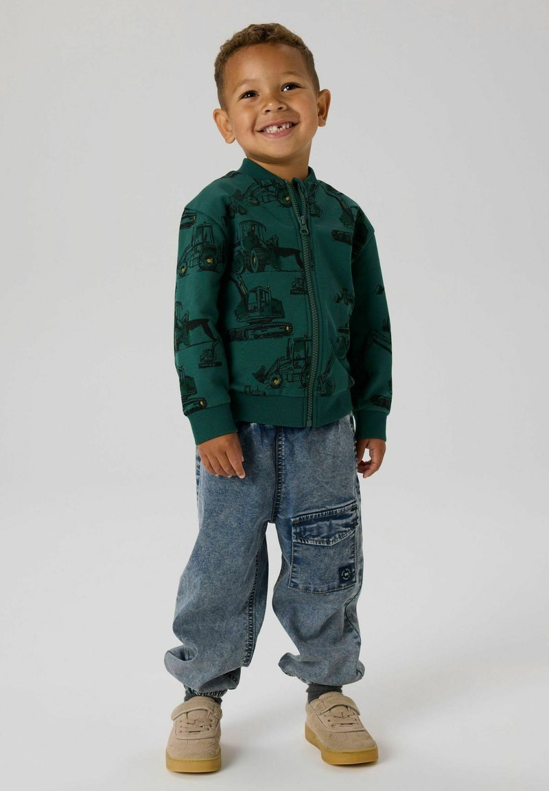 Zelená zipová bunda s potiskem černých stavebních vozidel, kombinovaná s volnými modrými džínami s velkou boční kapsou a šedými ponožkami.