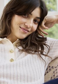 Giovane donna con lunghi capelli castani, indossa un maglione bianco a coste con bottoni dorati, appoggiando la testa sulla mano e sorridendo dolcemente.