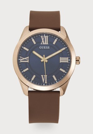 Montre avec bracelet en silicone marron, cadran bleu marine, accents en or rose, marqueurs en chiffres romains et aiguilles métalliques. Présente un boîtier rond et une couronne.