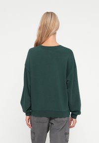 Felpa oversize verde scuro con scollo rotondo e spalle scese. Tessuto morbido, dettagli a costine sui polsini e sull'orlo.
