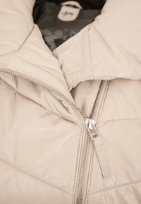 Beige Lederjacke mit gep quilteter Textur, ausgestattet mit einem silbernen Reißverschluss und einem Etikett, das im Kragen sichtbar ist.