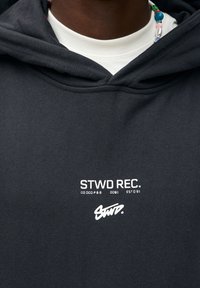 Czarna bawełniana bluza z kapturem z przednim logo w białym kolorze, z widoczną warstwową białą koszulą przy dekolcie oraz teksturowaną tkaniną.