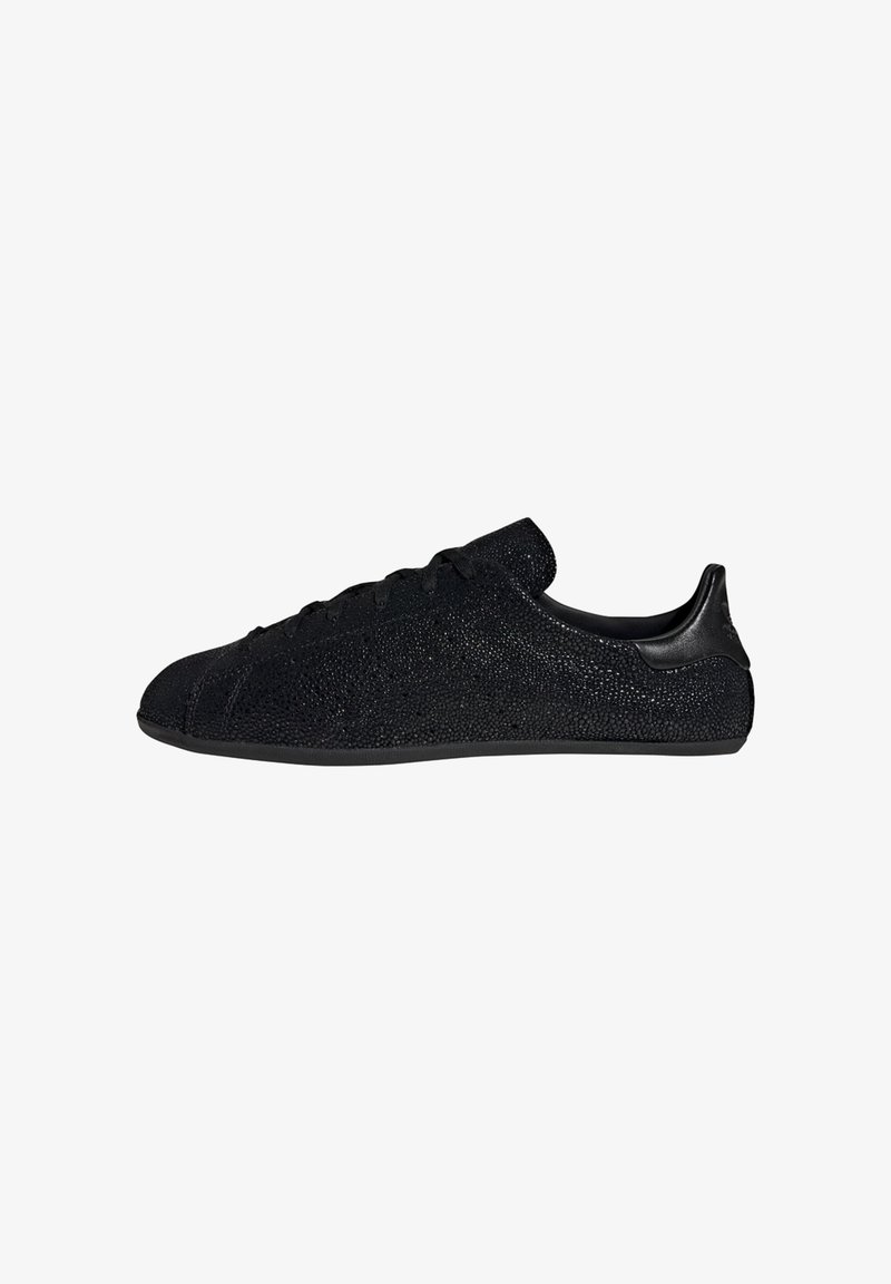 Μαύρο sneaker χαμηλού προφίλ με υφή στην επιφάνεια, σχέδιο με κορδόνια και λείο ταμπ ελάτης, απεικονισμένο σε προφίλ στο πλάι σε λευκό φόντο.