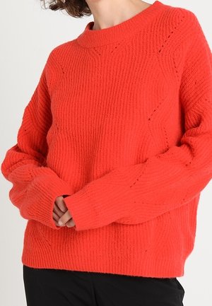 Frau trägt einen leuchtend orangefarbenen, strukturierten Strickpullover mit langen Ärmeln, kombiniert mit schwarzen Hosen vor neutralem Hintergrund.