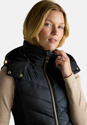 Blonde woman wearing a black quilted vest with gold zipper and buttons, beige long-sleeve top, holding the vest collar with her right hand.