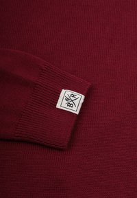 Burgundy stickad tröja med ribbad mudd och en vit tygetikett som visar en logotyp. Mjuk textur med ett enhetligt mönster.