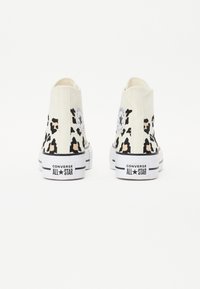 Vita högstövlar av sneakers med ett leopardmönster, featuring a cirkulär logotyp-patch och svart gummisula. Djärv text på hälen: "CONVERSE ALL STAR."