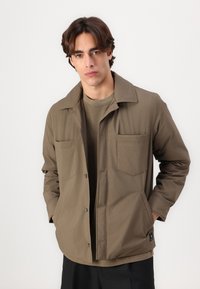 Brown lightweight jacket with a collared design, two chest pockets, and a front button closure. Worn over a matching olive green top.