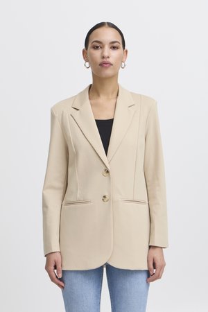 Kvinde med glat hår trukket tilbage iført guldringøreringe, beige blazer med to knapper over sort top og lyseblå jeans mod hvid baggrund.