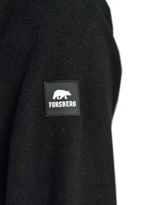 Zwart fleece-stof met een vierkant zwart etiket met een berenlogo en de tekst "FORSBERG." Textuur oppervlak, geen andere zichtbare patronen.