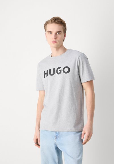 Сива памучна тениска с къси ръкави, с голям черен текст "HUGO" отпред, кръгло деколте и свободна кройка.