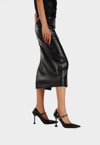 Le décolleté neri con tacco alto hanno una punta affusolata, pelle texturizzata e una cinghia alla caviglia con un accento in catena dorata. Indossati con una gonna in pelle nera.