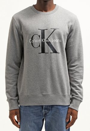 Man draagt een grijze Calvin Klein Jeans sweatshirt met zwart-witte "CK"-logo, gecombineerd met blauwe spijkerbroek.