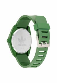 Žalias gumos dirželio laikrodis su nerūdijančio plieno nugarėle, turintis apvalią korpuso formą ir įpjovimo dizainą. Rodo Adidas logotipą.