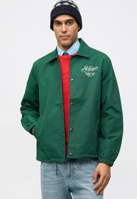 Άνδρας που φοράει μαύρο και άσπρο πλεκτό σκουφάκι, πράσινο μπουφάν με λογότυπο "Hilfiger Racing Club", κόκκινο πουλόβερ, ανοιχτό μπλε πουκάμισο και ανοιχτό μπλε τζιν.