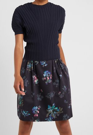 Personne portant un haut côtelé bleu marine à manches courtes et une jupe noire avec des motifs floraux colorés sur un fond clair uni.