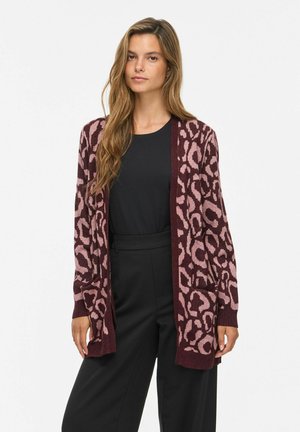 Femme portant un long cardigan ouvert avec un imprimé animal violet et rose sur un top noir et un pantalon noir, debout devant un fond blanc.