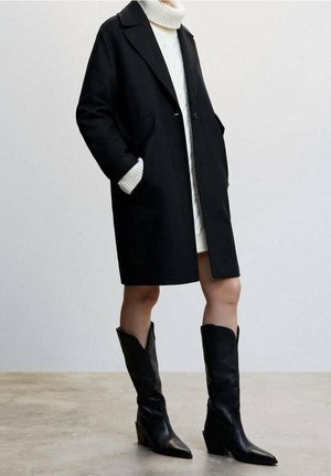 Personne portant un manteau noir longueur genoux sur une robe en tricot blanche, associée à des bottes noires à bout pointu mi-mollet, debout sur un sol neutre.