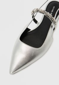 Ασημί μεταλλικό παπούτσι με μυτερή μύτη και σχέδιο με κοψίματα, με μια λουρίδα με καρφιά πάνω από το επάνω μέρος και μαύρη εσωτερική σόλα.