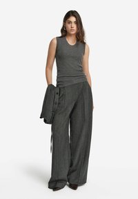 Kvinna iklädd en ärmlös grå ribbad topp och vida mönstrade byxor, hållande en matchande knäppt jacka i ena handen.