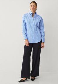 Blau-weiß gestreiftes Hemd mit Knopfleiste, Kragen und langen Ärmeln, kombiniert mit weit geschnittenen schwarzen Hosen und schwarzen spitzen Schuhen.