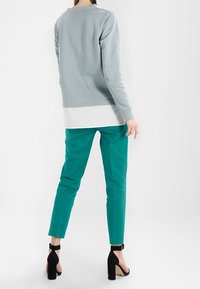 Suéter de manga larga gris claro superpuesto a una camisa blanca, combinado con pantalones de tela ajustados en color teal y sandalias de tacón negras.
