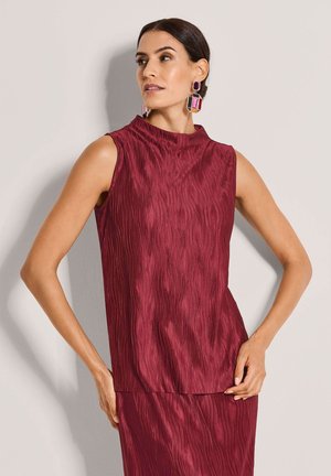 Vrouw die een mouwloze, gestructureerde bordeauxrode top en bijpassende rok draagt met grote geometrische oorbellen in meerdere kleuren, poserend tegen een effen achtergrond.