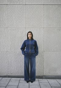 Trendi traper jakna i široke hlače u tamnoplavoj boji, s zatvaračem na zip i elastičnim manžetama, nosi se uz pozadinu od teksturiranog zida.