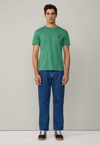 Camiseta de algodón verde con cuello redondo y mangas cortas, con detalles azules y un número "2" impreso en el pecho, combinada con vaqueros azules y zapatillas.